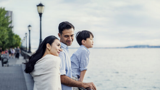 A woman, man, and child sit stand against a railing looking at the Hudson river with green trees behind them.